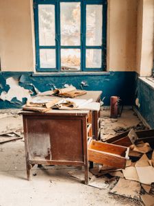 A deserted classroom showing an old desk and scattered papers in a dilapidated setting.